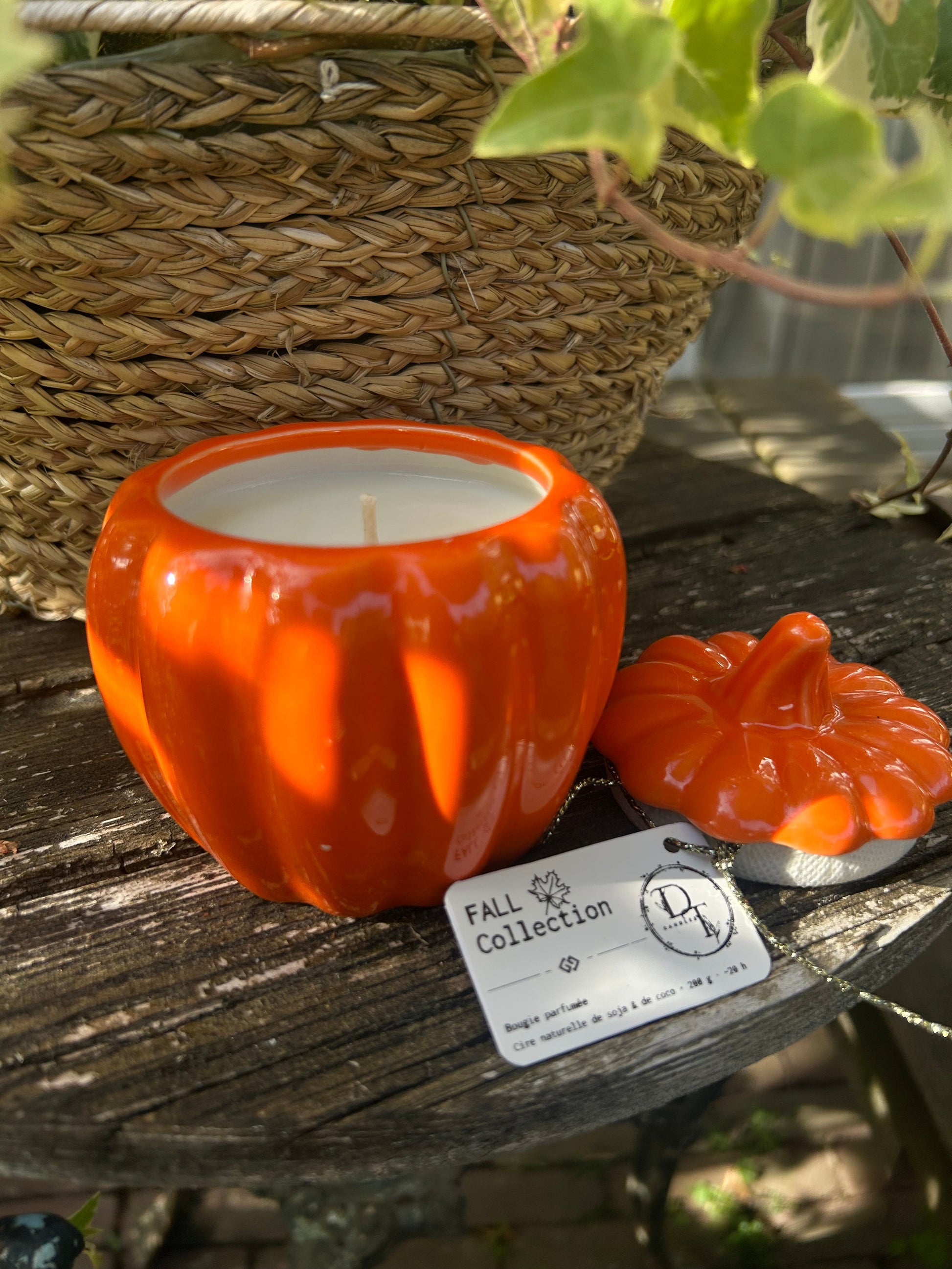 Orange pumpkin-shaped candle with a label on a wooden surface, surrounded by greenery.