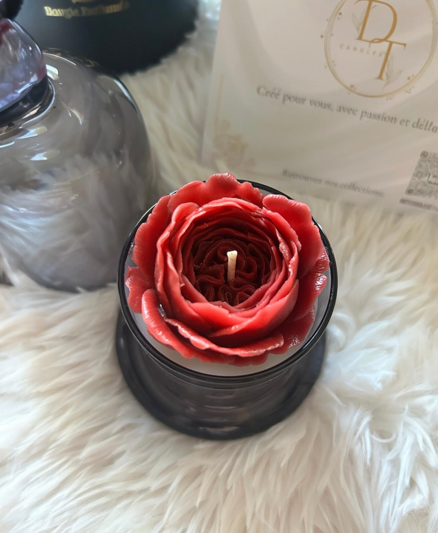 Red rose-shaped candle in a black holder on a fluffy white surface with a brand name visible in the background.