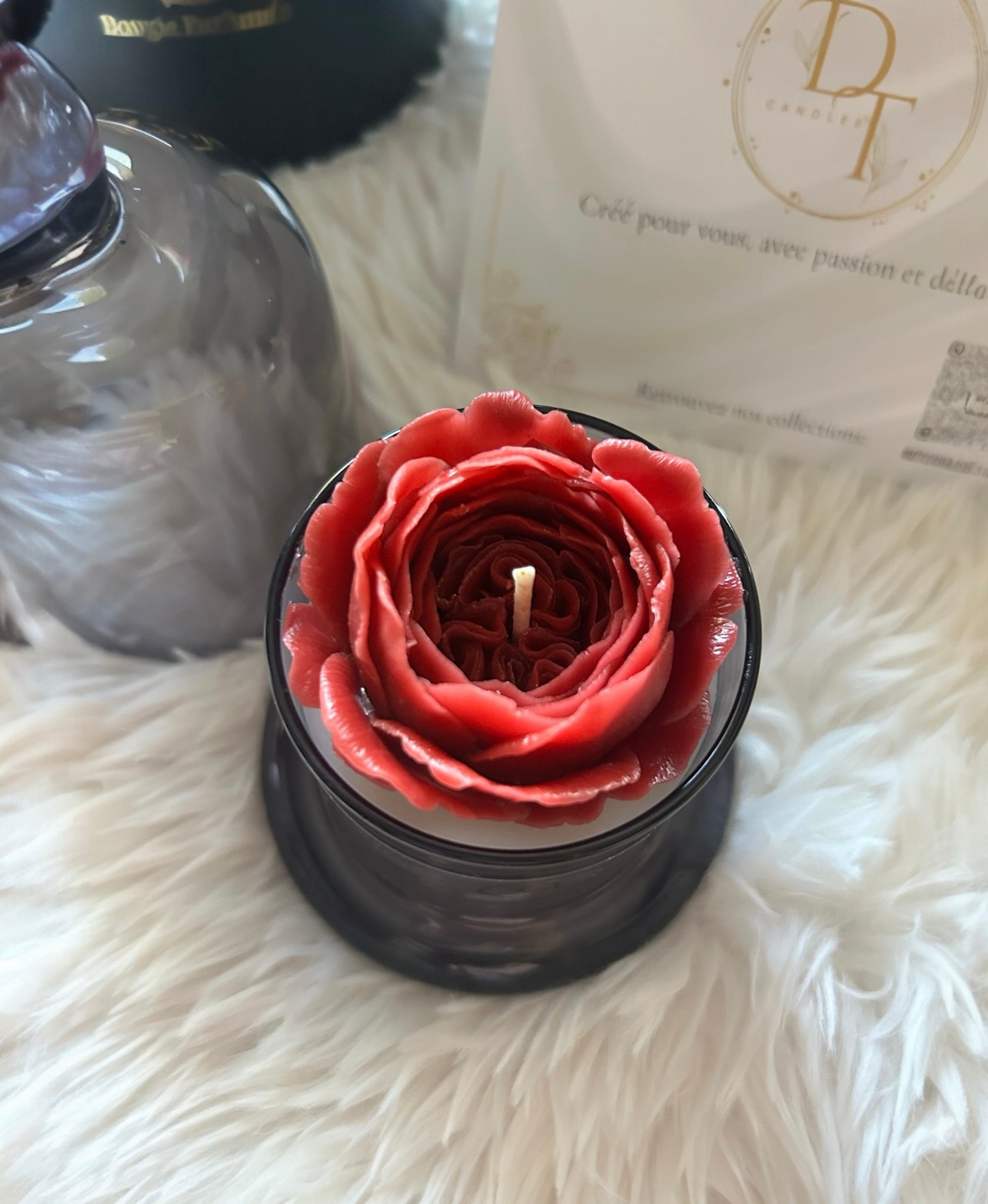 Red rose-shaped candle in a black holder on a fluffy white surface with a brand name visible in the background.