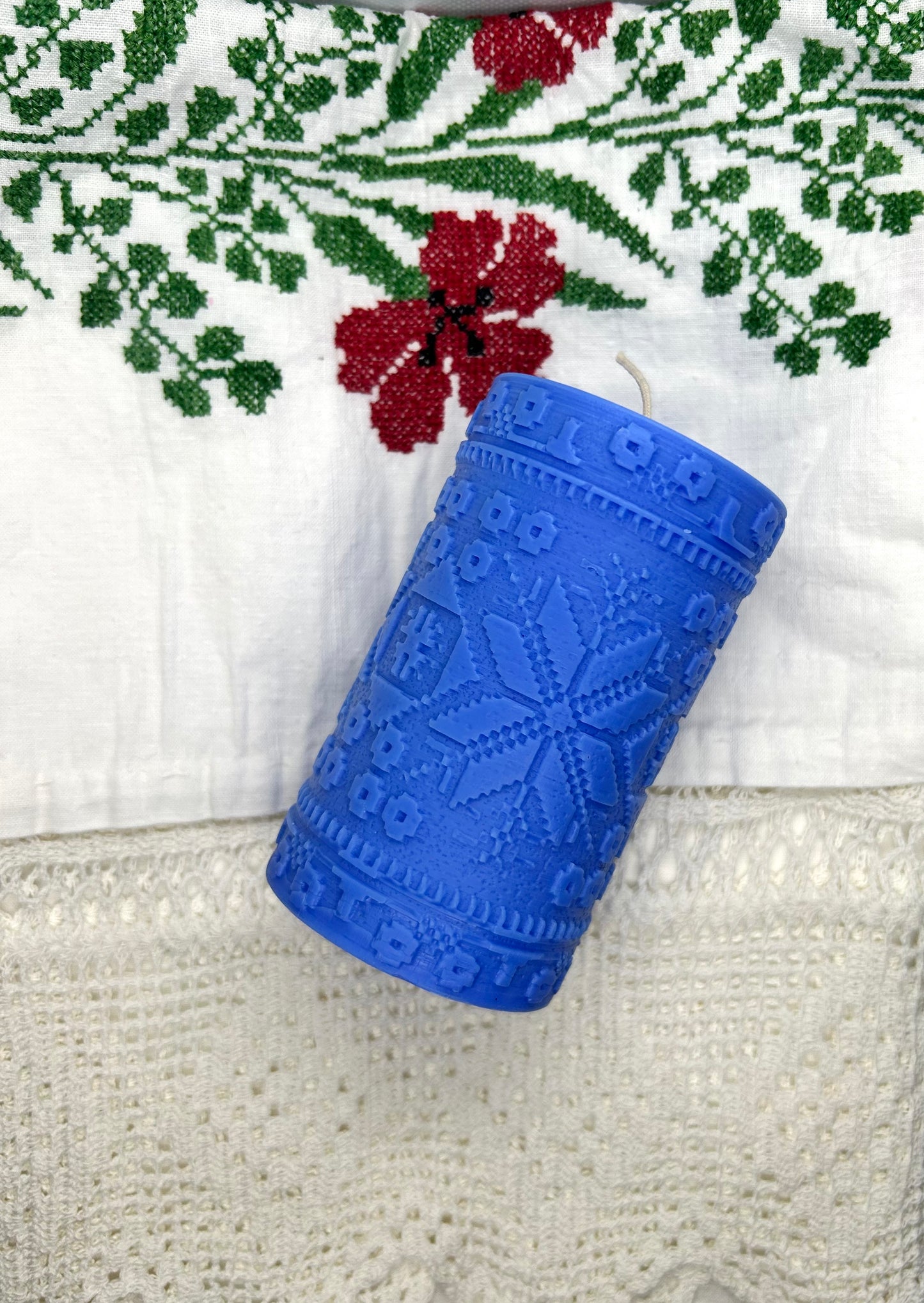 A blue handcrafted scented candle with an embossed ethnic-inspired motif, placed on a white textured surface with a floral patterned fabric in the background.