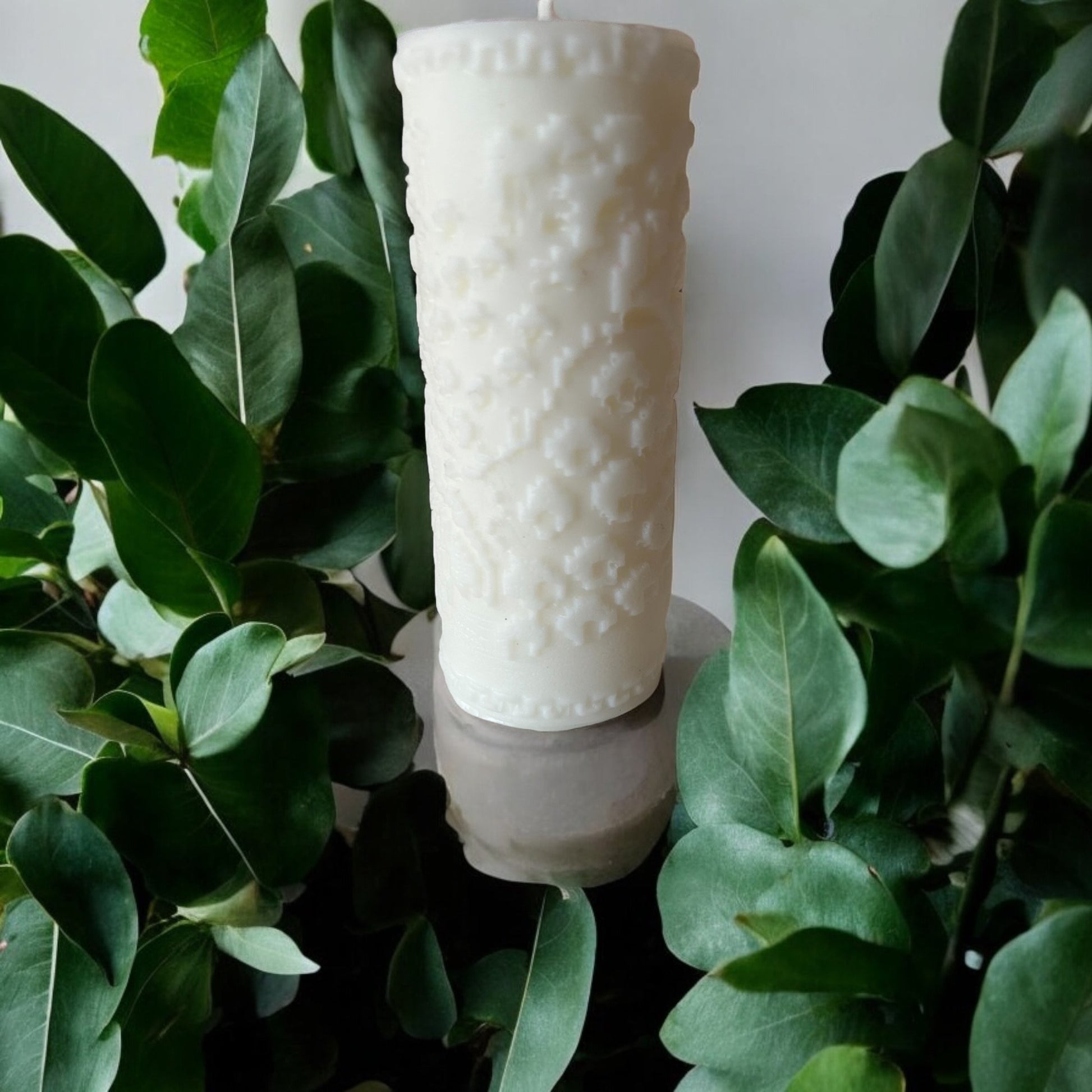 A white, handcrafted scented candle with intricate embossed patterns, displayed amidst green leaves.